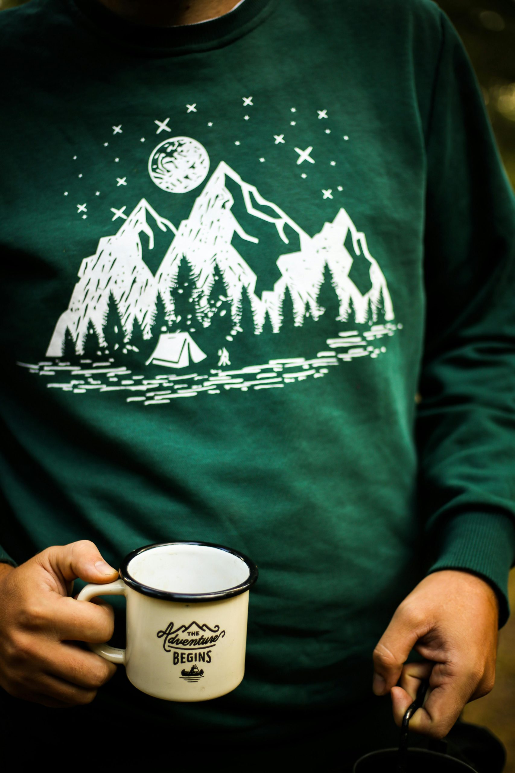 Person holding a mug with outdoor adventure design on a green sweater background.